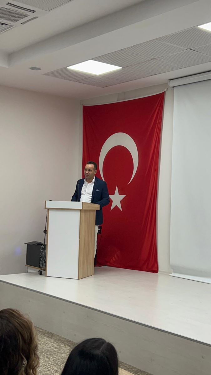 Kürsüde konuşan bir kişi; arka planda duvara asılı büyük Türk bayrağı, sağda projeksiyon perdesi olan salon.