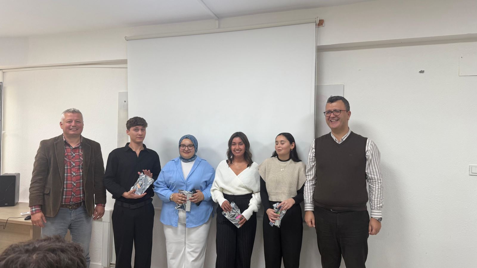 Bir sınıf ortamında, projeksiyon perdesi önünde yan yana duran iki öğretim elemanı ile dört öğrenci görülüyor. Öğrenciler ellerinde küçük hediye paketleri tutuyor. Herkes kameraya doğru gülümsüyor.