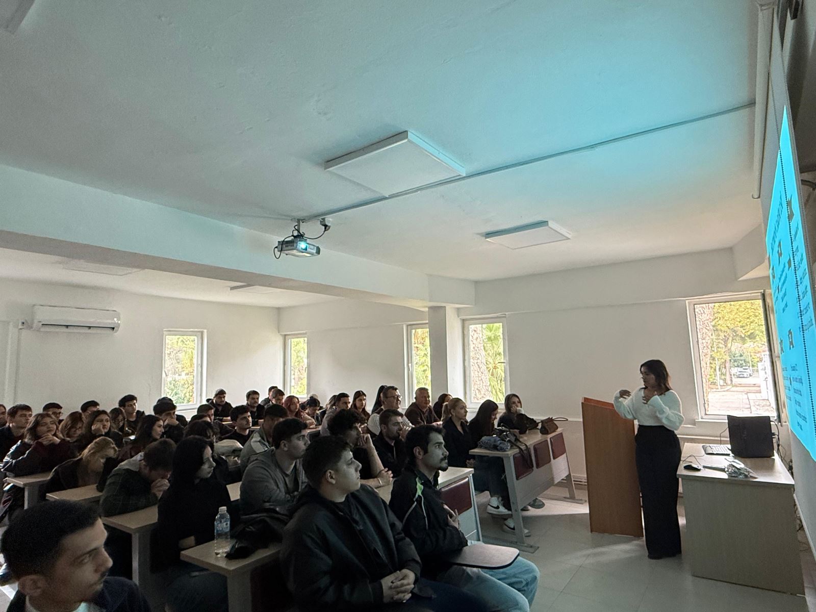Sınıfın önünde duran bir öğrenci sunum yaparken, sıralarda oturan çok sayıda öğrenci sunumu izliyor. Projeksiyon perdesinde lojistikle ilgili grafik ve maddeler içeren bir slayt görüntüleniyor.