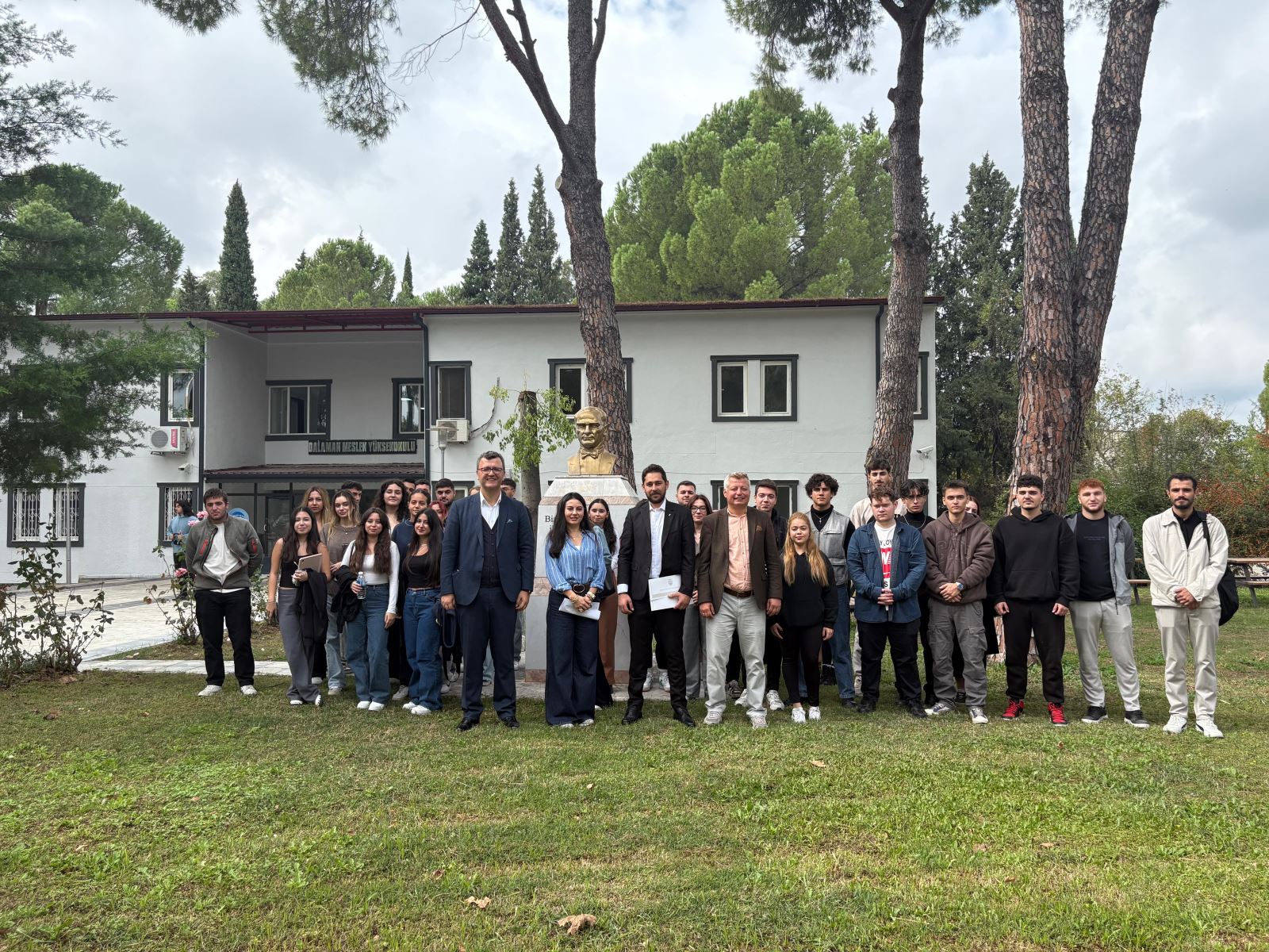 "Dalaman MYO dersliğinde Mezun Öğrenci Buluşması konferansı. Arkada Atatürk portresi ve projeksiyon perdesi. Kudret Hasgül sunum yaparken öğrenciler ve akademisyenler dikkatle dinliyor. 23 Ekim 2025 tarihli etkinlik görüntüsü."