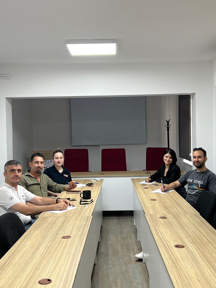 The photograph captures five people sitting around a long table in a modern, minimalist meeting room, all smiling and looking directly at the camera.  The room features light gray flooring and plain white walls. The tables have a light-colored wood-grain surface with white bases, arranged in two long rows that form a "U" shape, leaving a wide aisle down the middle.
