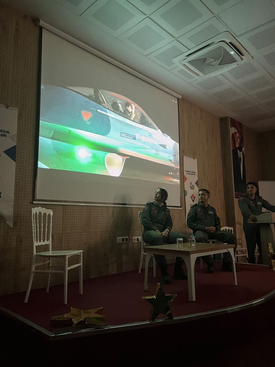 Bir konferans salonunun sahnesinde, yeşil uçuş tulumu giymiş askeri personeller bulunuyor. Sahnedeki büyük projeksiyon perdesinde bir savaş uçağının görüntüsü yansıtılıyor. Sahnenin ortasında oturan biri kadın iki askeri personel perdeye bakarken, sağ taraftaki kürsüde bir diğer askeri personel ayakta duruyor. Sahne önünde altın renkli yıldız dekorları yer alıyor.  