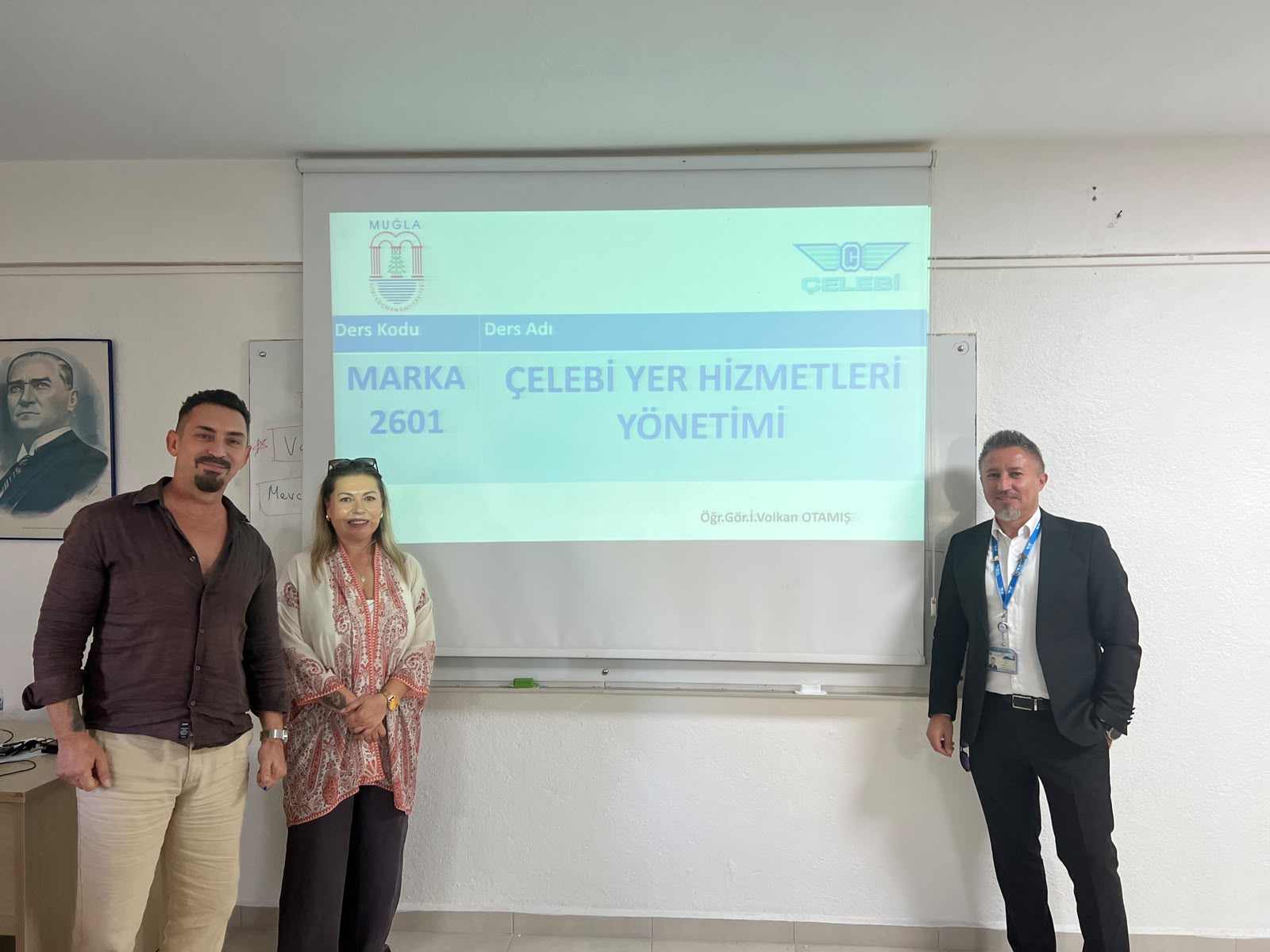 Bir üniversite dersliğinde projeksiyon perdesinin önünde duran üç kişi. En solda kahverengi gömlekli ve bej pantolonlu bir erkek, ortada desenli hırka ve koyu renk pantolonlu bir kadın, sağda ise siyah takım elbiseli, boynunda yaka kartı taşıyan bir erkek bulunuyor. Arka plandaki projeksiyon perdesinde "Muğla Sıtkı Koçman Üniversitesi" ve "Çelebi Hava Servisi" logoları ile "MARKA 2601 - ÇELEBİ YER HİZMETLERİ YÖNETİMİ" yazısı görünmektedir.  