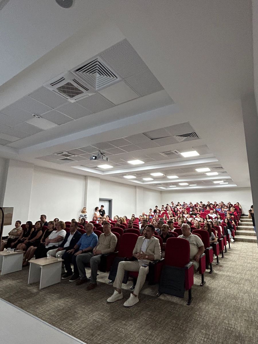 This shot was taken in a lecture hall. The hall is spacious, with white walls and square-shaped ceiling lights. Rows of red chairs rise upwards. In the front rows sit faculty members in suits and simple attire; most are solemnly but attentively gazing toward the stage. The back rows are filled with students. A projector hangs from the ceiling, and a presentation is taking place. The atmosphere is formal yet informal