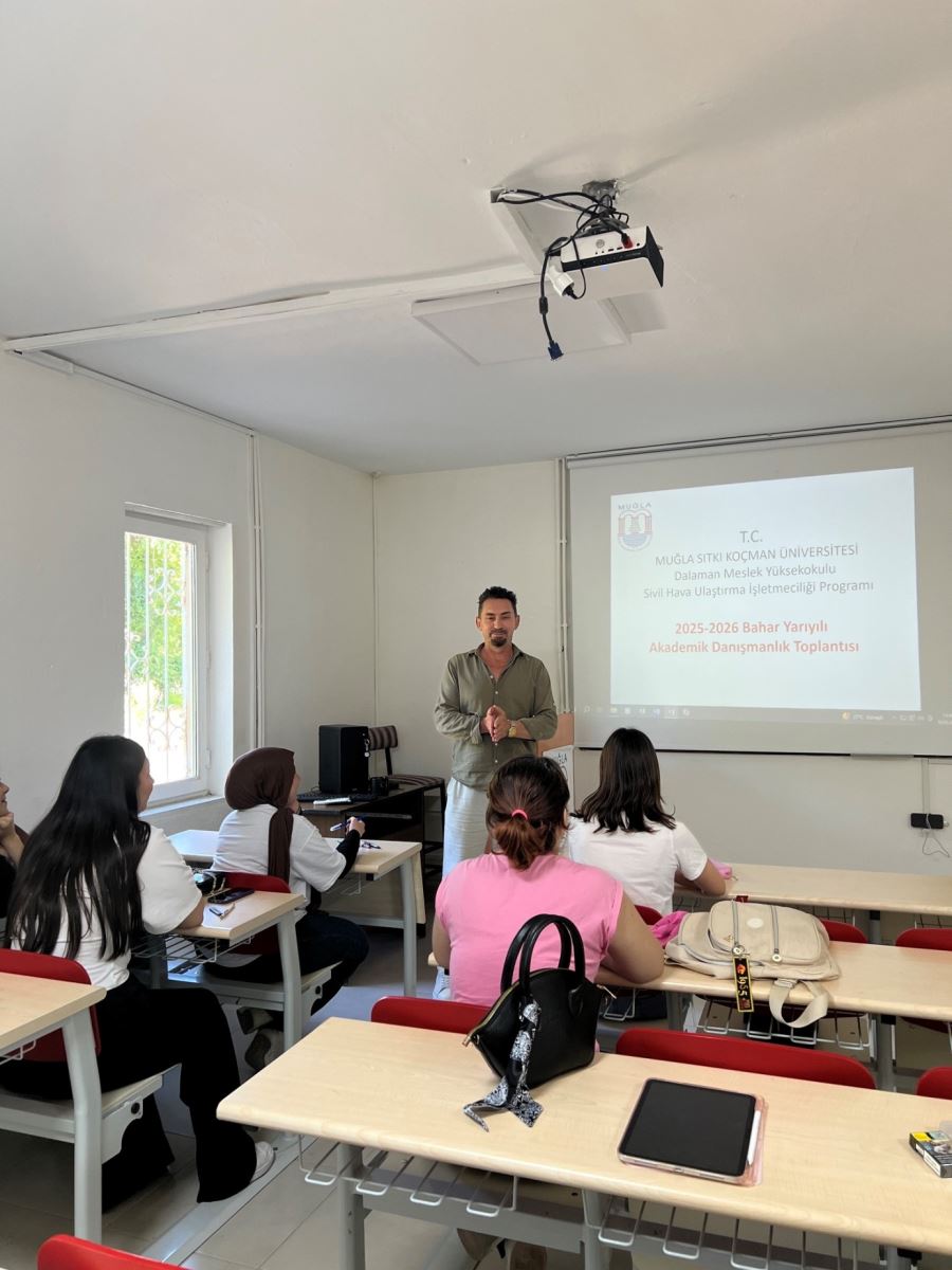 An academic meeting taking place inside a university classroom.  Background and Setting: A bright classroom with white walls. A projector is mounted on the ceiling. On the right wall, there is a projection screen displaying the following Turkish text: "T.C. MUĞLA SITKI KOÇMAN ÜNİVERSİTESİ, Dalaman Meslek Yüksekokulu, Sivil Hava Ulaştırma İşletmeciliği Programı, 2025-2026 Bahar Yarıyılı Akademik Danışmanlık Toplantısı" (Muğla Sıtkı Koçman University, Dalaman Vocational School, Civil Aviation Transportation Management Program, 2025-2026 Spring Semester Academic Counseling Meeting). On the left, there is a vertical window with iron bars showing greenery outside.  Main Subject (Instructor): Standing at the front of the classroom, just to the left of the projection screen, is a male instructor in his 30s or 40s with dark hair, a light beard, and a mustache. He is wearing a khaki green shirt and white trousers. His hands are clasped together in front of him as he addresses the class.  Students: Four female students are seated in the front rows, viewed from behind. The student on the far left and the one next to her wearing a hijab are both in white t-shirts. In the middle, there is a student in a pink blouse, and to her right is another student in a white t-shirt. They are all facing toward the instructor and the screen.  Foreground Details: On the empty desk in the immediate foreground, there is a black handbag, a tablet, and a black glasses case. On the adjacent desk, a cream-colored backpack is visible. The chairs in the classroom have red backrests.