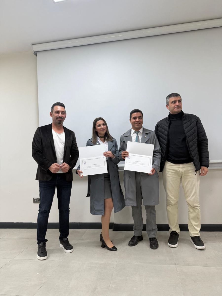 This photograph shows four individuals standing side-by-side in front of a white wall and a white projection screen. The man and woman in the center are wearing long, grey/blue trench coat-like outerwear and are both holding rectangular, white certificates. The man on the far left is wearing a black velvet jacket, a white t-shirt, and dark jeans. The man on the far right is wearing a black puffer jacket, a black turtleneck, and light beige pants. They are all looking at the camera and smiling. The setting appears to be a classroom or meeting room.