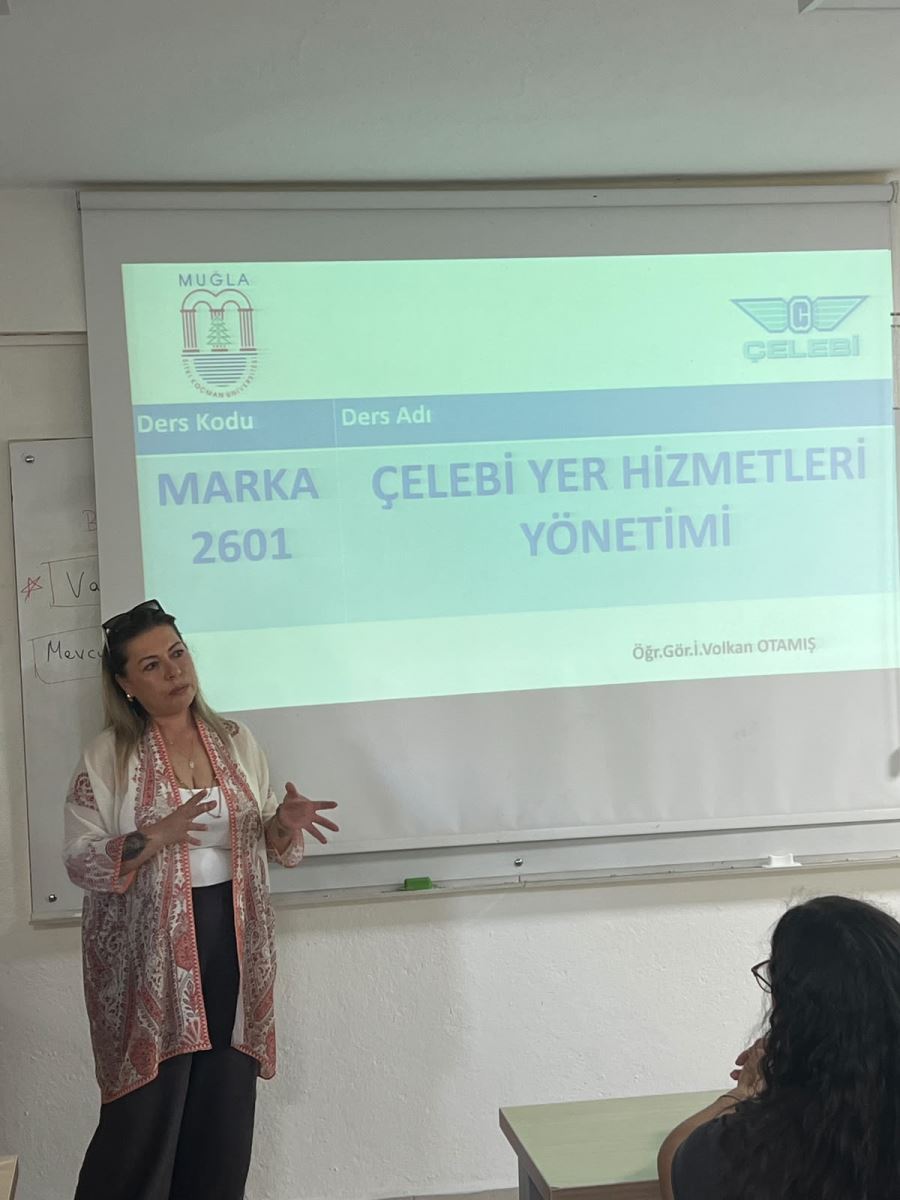 Derslikteki projeksiyon perdesinin sol ön tarafında duran kadın konuşmacı. Desenli hırkası ve siyah pantolonuyla ellerini açıklama yaparken hareket ettiriyor. Arka plandaki perdede dersin sunum slaytı açık. Ön planda sağ altta bir öğrencinin başı ve omzu görünmektedir.  