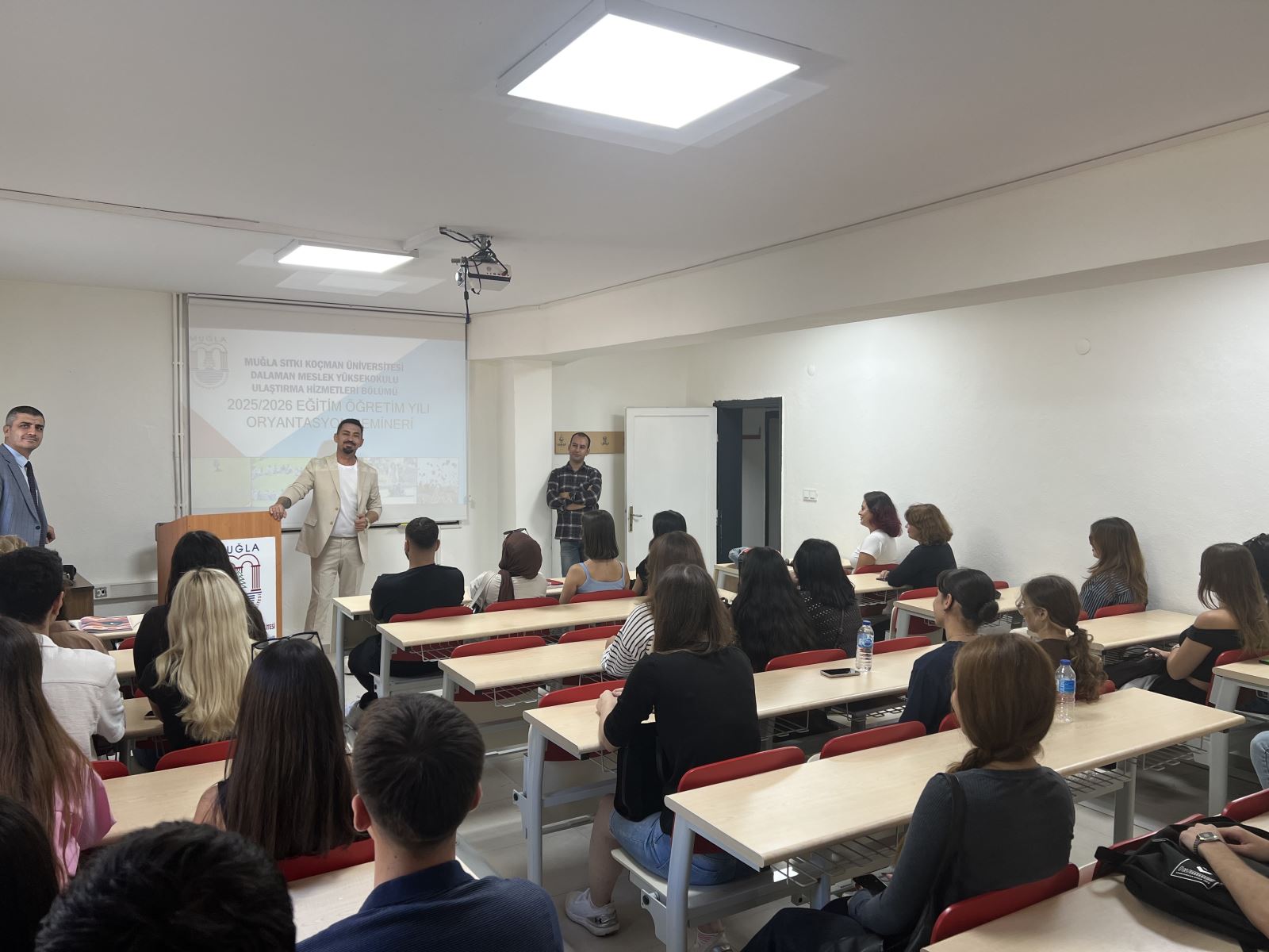 This photo was taken in a classroom. In the foreground, young students are seated at desks; most are women, a few are men. All are looking intently at the blackboard or the speaker. The Department Head is giving a speech in the foreground; he's wearing a light-colored suit and supporting his explanation with hand gestures. In the background is a projection screen with the following text: "Muğla Sıtkı Koçman University, Dalaman Vocational School, Department of Transportation Services, 2025/2026 Academic Year Orientation Seminar."  Next to the speaker are two other faculty members from the department; one is wearing a suit, the other a shirt. The classroom atmosphere is orderly, bright, and welcoming.
