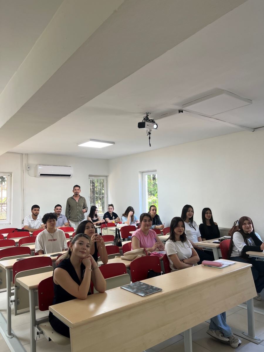 A wide-angle shot of the same classroom, this time taken from the front/side angle, showing the faces of the students.  Background and Setting: A white air conditioner is mounted on the back wall of the classroom. Another vertical window is visible on the left side. A rectangular LED panel light and the projector on the ceiling are clearly visible. Rows of wooden desks with red-backed chairs fill the room.  Students and Instructor: There are approximately 13 to 14 students in the classroom. The male instructor in the khaki shirt from the first photo is now standing at the very back of the room among the students. Both male and female students are seated at the desks.  Foreground Details: In the very front row, closest to the camera, a young female student wearing a black sleeveless top rests her chin on her hands, smiling directly at the camera. To her right, another female student in a white t-shirt sits with her arms crossed, looking slightly past the camera with a gentle smile. The rest of the students in the background are seated and looking forward attentively. Notebooks, pencil cases, and pens are scattered on the desks.