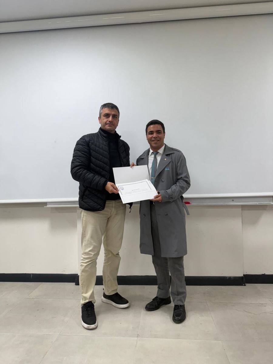 This vertical photograph features two men standing in front of a white projection screen. The man on the left, wearing the black puffer jacket and beige pants, is presenting a certificate to, or holding the certificate with, the man on the right, who is wearing the grey trench coat. Both are looking at the camera.