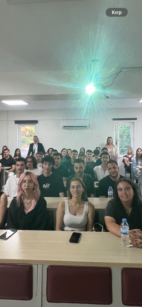 Sınıftaki öğrencilerin sıradan arkaya doğru dizildiği cephe açısı. Ön sıralarda oturan üniversite öğrencileri doğrudan kameraya bakıyor. Arka tarafta ayakta duran ve dersi takip eden eğitmenler görünmektedir. Tavandaki projeksiyon cihazından güçlü bir ışık yansımaktadır.  