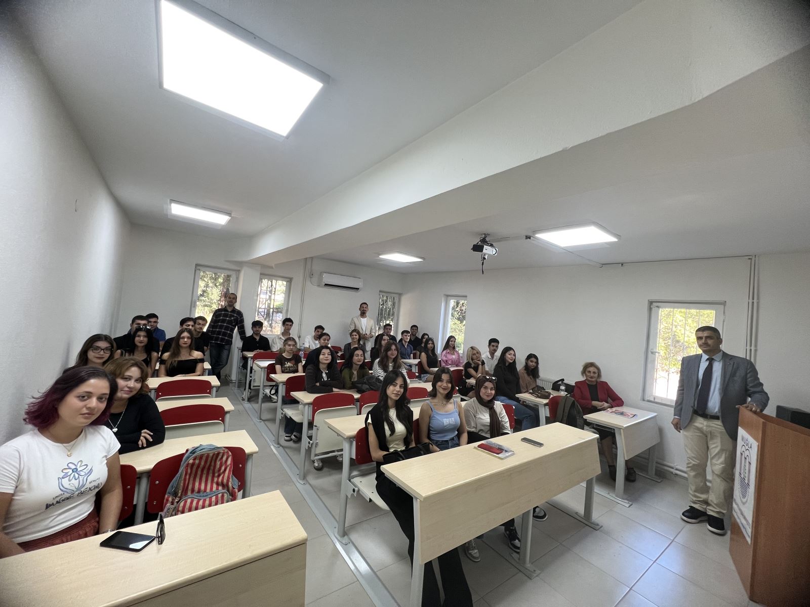 This photo was taken in a classroom. Students are sitting at desks, three male faculty members are standing between them, and they all smile at the photographer at the front of the room.