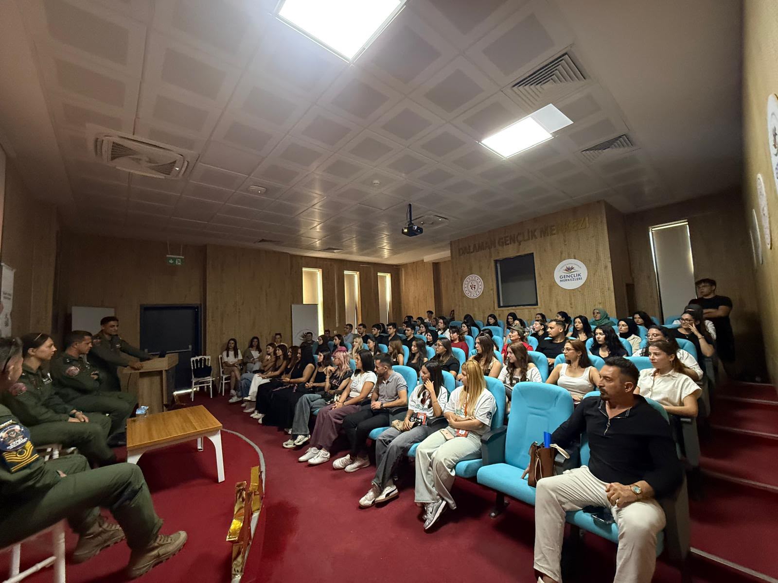 Konferans salonunun arka sağ köşesinden sahneye ve dinleyicilere doğru çekilmiş açı. Sağ ön sırada siyah tulum/gömlek ve açık renk pantolonlu bir erkek oturuyor. Mavi koltuklarda oturan öğrenciler, sol taraftaki sahnede yer alan askeri personellerin sunumunu takip ediyor.  