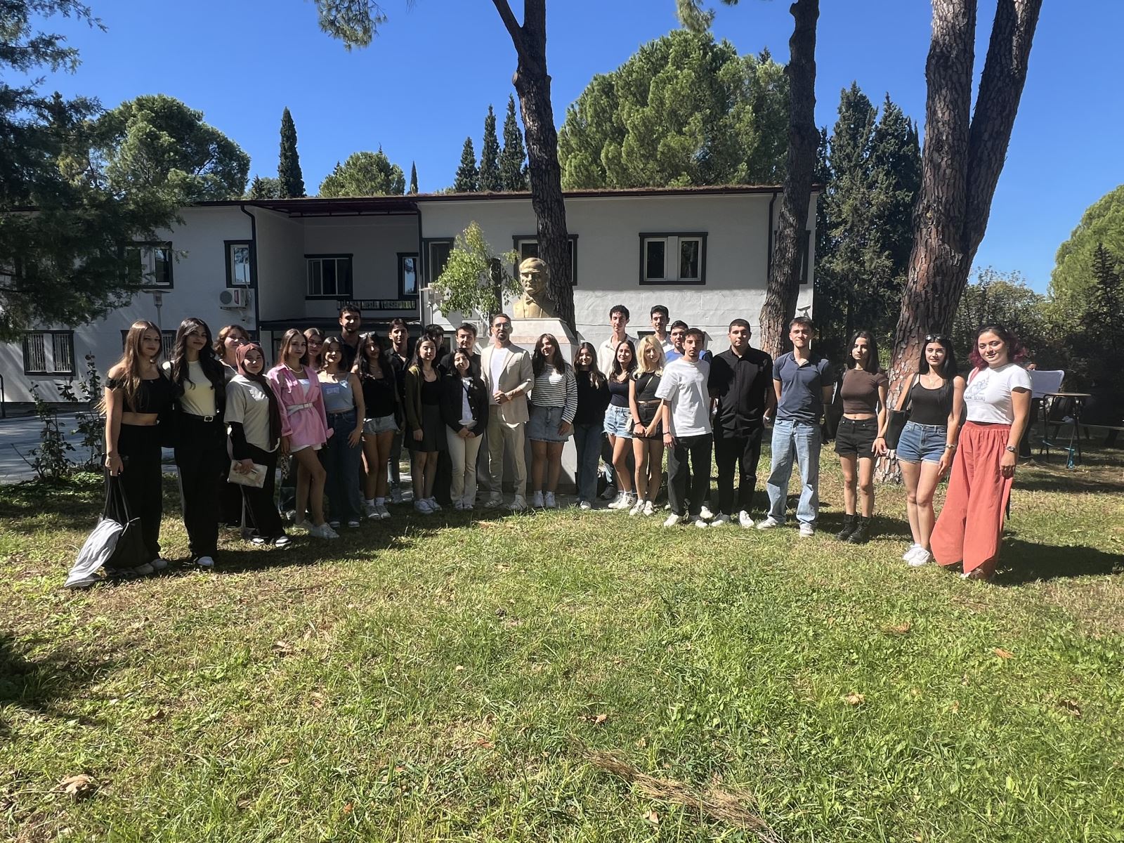 This photo was taken outdoors on a sunny day. A large group of students are standing together on green grass. In the background is a white building with a bust of Atatürk in front of it. There are approximately 25 people in the photo; most are young and dressed in casual summer clothes. There are also a few men.  In the center is a lecturer in a light-colored suit. Everyone is smiling at the camera; the atmosphere is lively, cheerful, and friendly, reminiscent of a school day