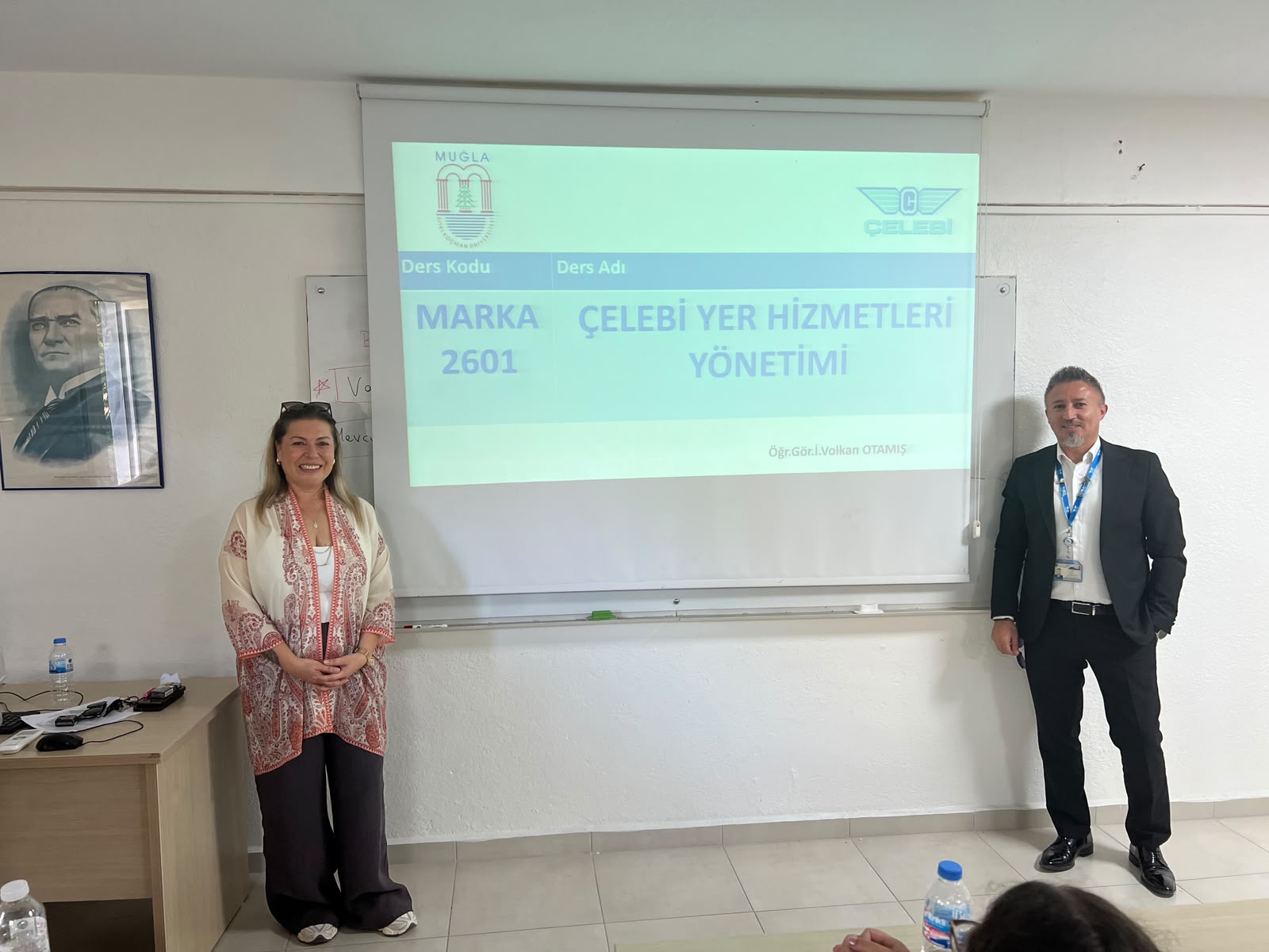 Projeksiyon perdesinin iki yanında duran iki kişi. Sol tarafta desenli hırkalı kadın konuşmacı, sağ tarafta siyah takım elbiseli ve yaka kartlı erkek konuşmacı kameraya gülümseyerek poz veriyor. Perdede ders adı ve logosu yer almaktadır.  