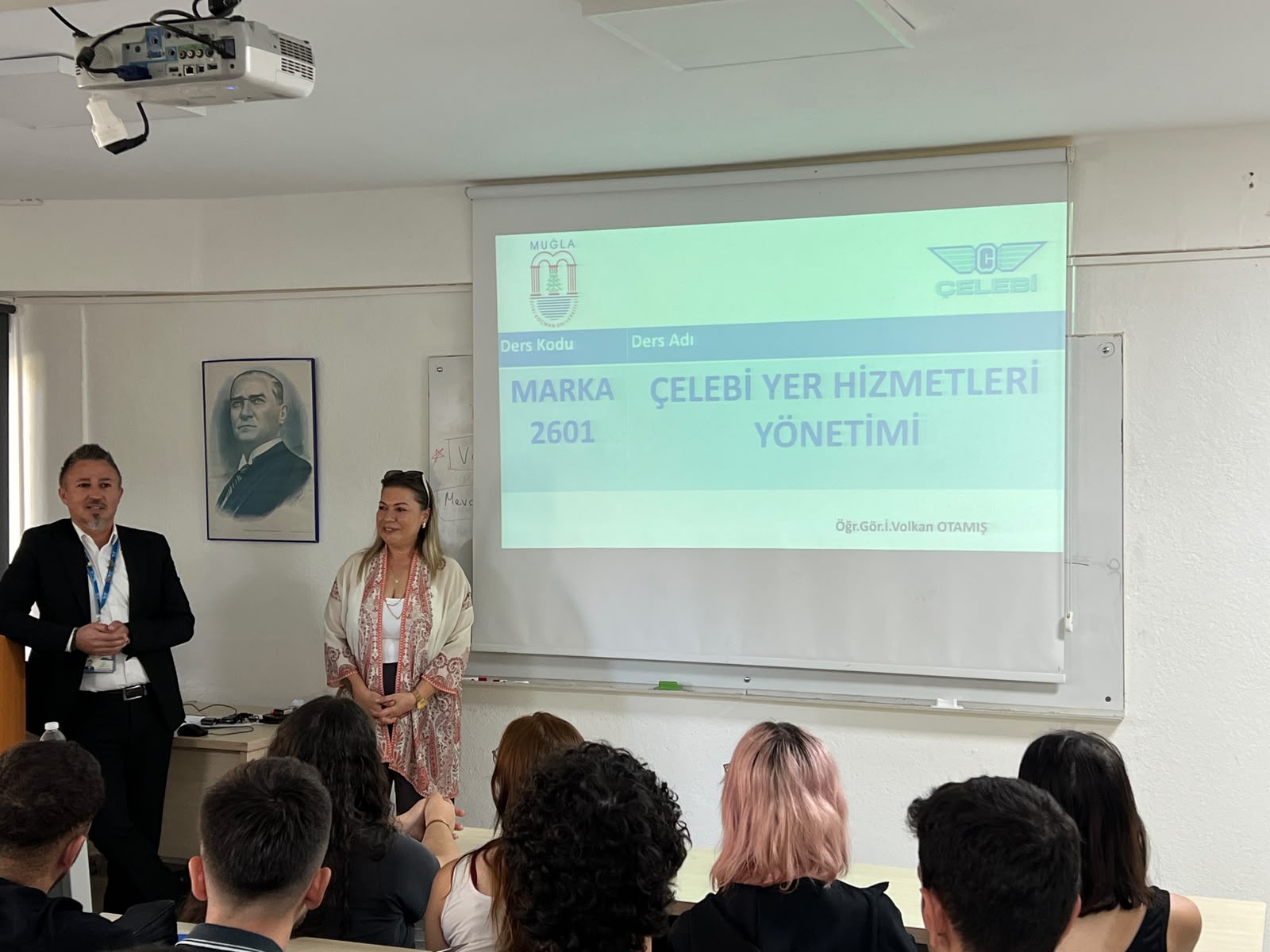 Ön sıralardaki öğrencilerin arkasından tahtaya doğru çekilmiş açı. Projeksiyon perdesinin solunda siyah takım elbiseli konuşmacı, sağında ise desenli hırkalı kadın konuşmacı ayakta durmaktadır. Duvarda Mustafa Kemal Atatürk'ün portresi asılıdır.  