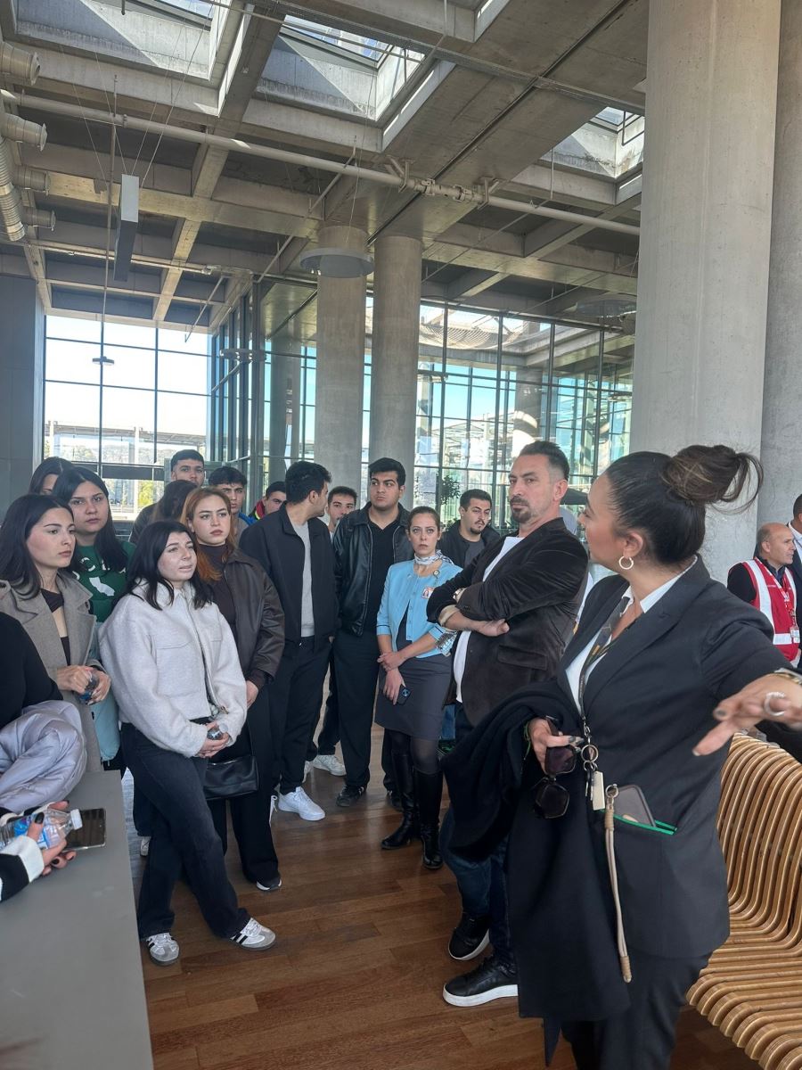 Terminal içindeki geniş beton sütunların arasında çekilmiş bir başka kare. Siyah ceketli kadın yetkili, grubu etrafında toplamış konuşmaya devam ediyor. Arka planda havalimanı yönlendirme tabelaları (32, 31A-31B gibi kapı numaraları) ve terminalin cam cephesinden içeri giren parlak gün ışığı görülüyor. Yerler ahşap parke döşeme.