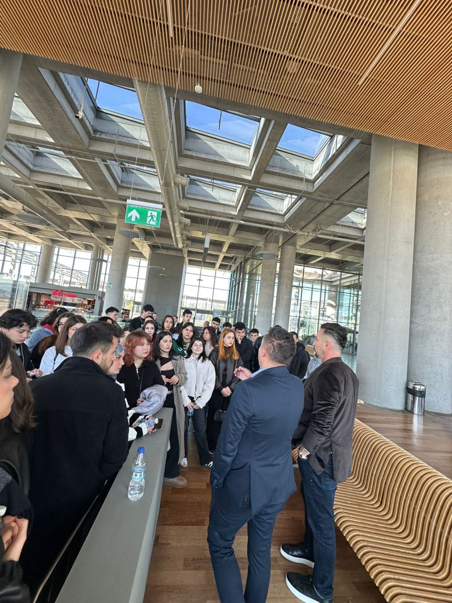 Önceki fotoğrafa benzer bir noktadan, ancak farklı bir açıyla çekilmiş. Bu karede, siyah ceketli bir kadın yetkilinin gruba bir şeyler anlatırken el hareketleriyle yönlendirme yaptığı görülüyor. Grubun arasında turkuaz renkli üniforması ve fularıyla bir Çelebi personeli de bulunuyor. Gençlerin yüzündeki odaklanmış ve ciddi ifade, teknik gezinin eğitici yanını vurguluyor..