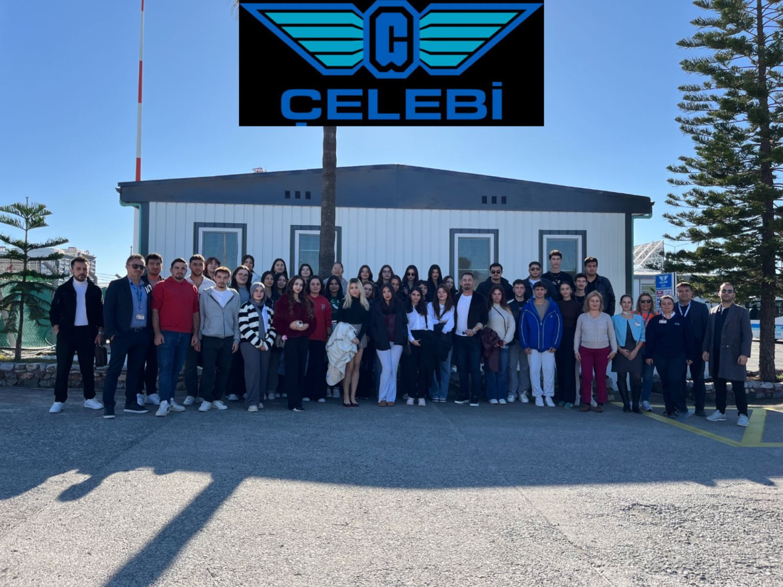 Grup bu kez üzerinde büyük bir mavi kanatlı logo ve "ÇELEBİ" yazısı olan beyaz, prefabrik tarzda bir binanın önünde. Binanın arkasında bir palmiye ağacı ve sağ tarafta yüksek çam ağaçları görülüyor. Zemin beton ve güneş ışığı tam karşıdan geldiği için fotoğrafta keskin gölgeler oluşmuş. Bu görsel, gezinin Çelebi Yer Hizmetleri ayağını temsil ediyor