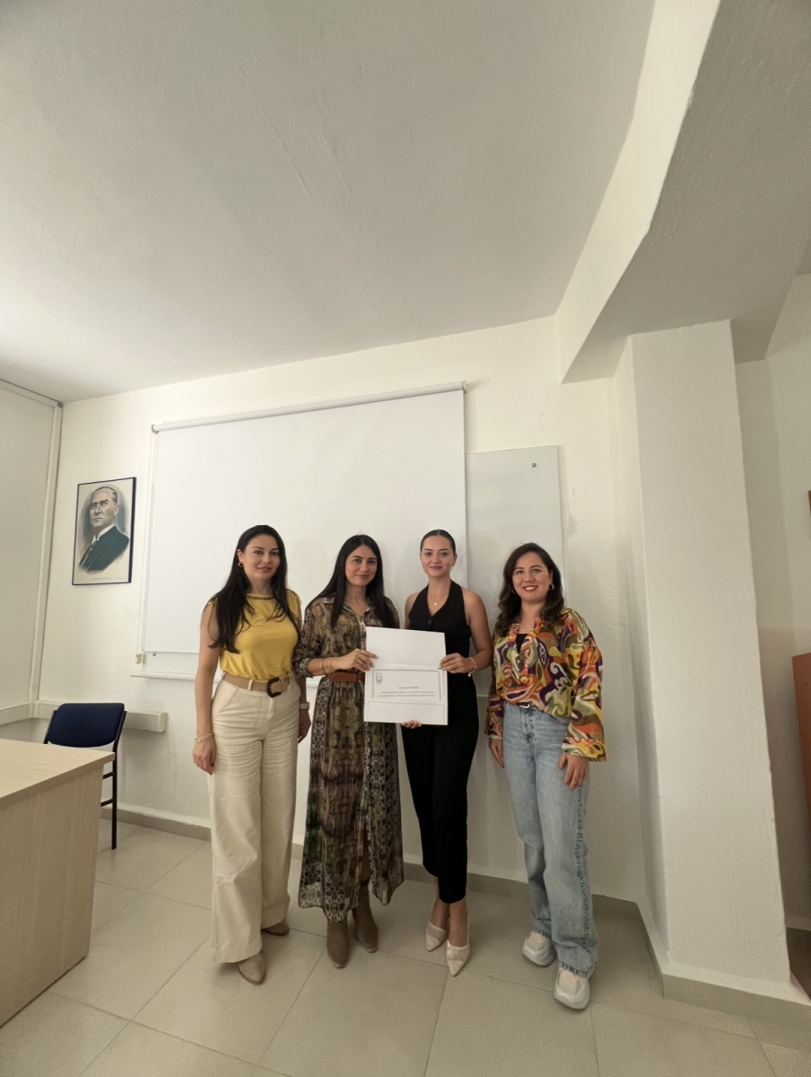 The photo is taken indoors, likely in a classroom or presentation room. In the background, there is a white screen (projection screen) and a whiteboard to its right. A framed portrait of Atatürk hangs in the upper left corner. The floor and walls are light-colored tiles and paint, giving the space a bright and neutral tone. Four young women are standing side by side, smiling at the camera. They are all dressed stylishly. The two women in the middle are holding a certificate together. •	The woman on the far left is wearing long, light beige pants and a sleeveless yellow blouse. She has dark, straight hair worn down. •	The second woman from the left is wearing a long patterned dress with brown-toned heeled boots. Her hair is also long, straight, and dark. •	The third woman (one of the certificate holders) is dressed in a black jumpsuit with white high heels. Her hair is tied back. •	The woman on the far right is wearing a colorful patterned blouse with light blue jeans and white sneakers. Her long, wavy dark hair is worn down.