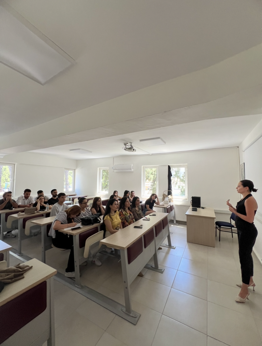 The photo is taken in a classroom setting. The environment and the people in the scene appear to be lit by natural daylight. The walls and floors are light-colored. Three large windows are located at the back of the classroom, and trees can be seen outside. The scene is as follows: On the right side, at the front of the class, a woman in a black jumpsuit is standing. She is wearing white high-heeled shoes, and her hair is tied back. She appears to be speaking or giving a presentation, holding an object in her hand. There is a computer and a chair beside her. On the left side, around 18–20 students are seated at desks. The majority are young women, though a few male students are visible in the back rows. All are attentively focused on the speaker. Most have bags, phones, or notebooks in front of them. The seating is arranged in three rows, typical of a university classroom. The atmosphere is organized, formal, and educational. The participants are dressed casually but neatly.