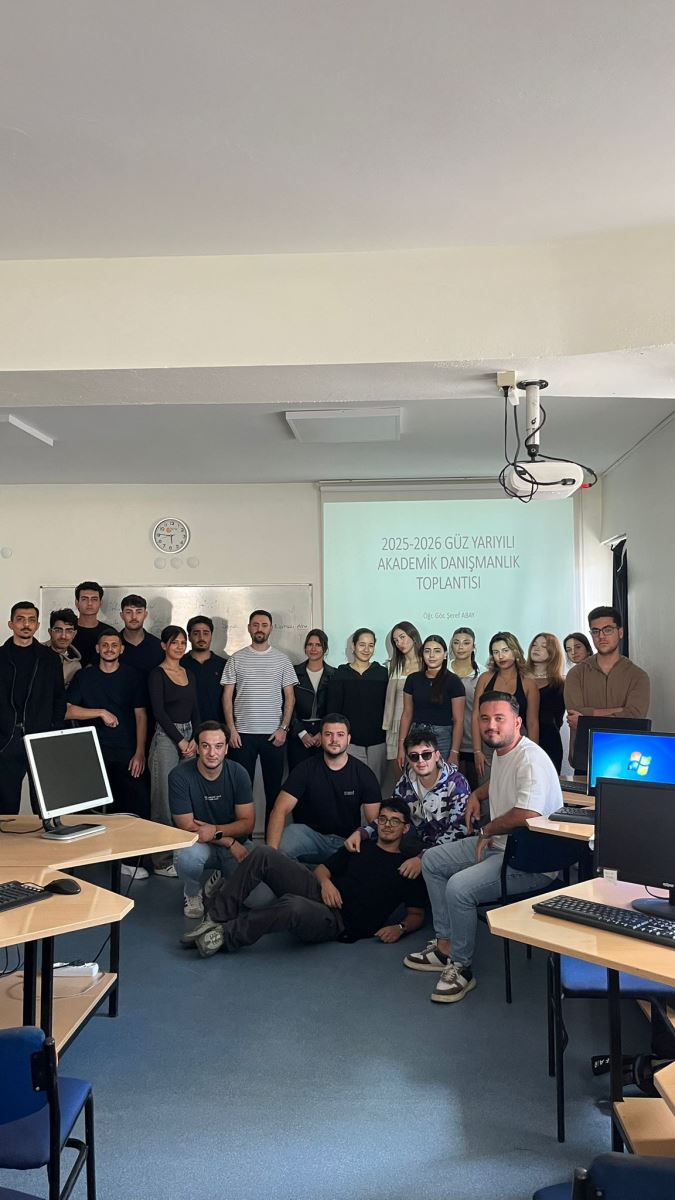  Bu fotoğrafta öğretim görevlisi tahtanın yanında ayakta durmaktadır.  Öğretim görevlisinin yanında ve önünde öğrenciler fotoğraf çekilmek için durmaktadır. Fotoğraf bilgisayar laboratuvarında çekilmiştir. Projeksiyon perdesine yansıtılmış beyaz arka planlı bir sunum slaytında şu yazı yer almaktadır: “2025-2026 GÜZ YARIYILI AKADEMİK DANIŞMANLIK TOPLANTISI Öğr. Gör. Şeref ABAY” Masanın üzerinde bilgisayarlar ve tavanın sağ tarafında projeksiyon cihazı vardır.  