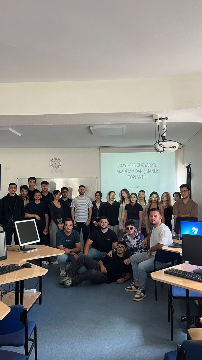  Bu fotoğrafta öğretim görevlisi tahtanın yanında ayakta durmaktadır.  Öğretim görevlisinin yanında ve önünde öğrenciler fotoğraf çekilmek için durmaktadır. Fotoğraf bilgisayar laboratuvarında çekilmiştir. Projeksiyon perdesine yansıtılmış beyaz arka planlı bir sunum slaytında şu yazı yer almaktadır: “2025-2026 GÜZ YARIYILI AKADEMİK DANIŞMANLIK TOPLANTISI Öğr. Gör. Şeref ABAY” Masanın üzerinde bilgisayarlar ve tavanın sağ tarafında projeksiyon cihazı vardır.  