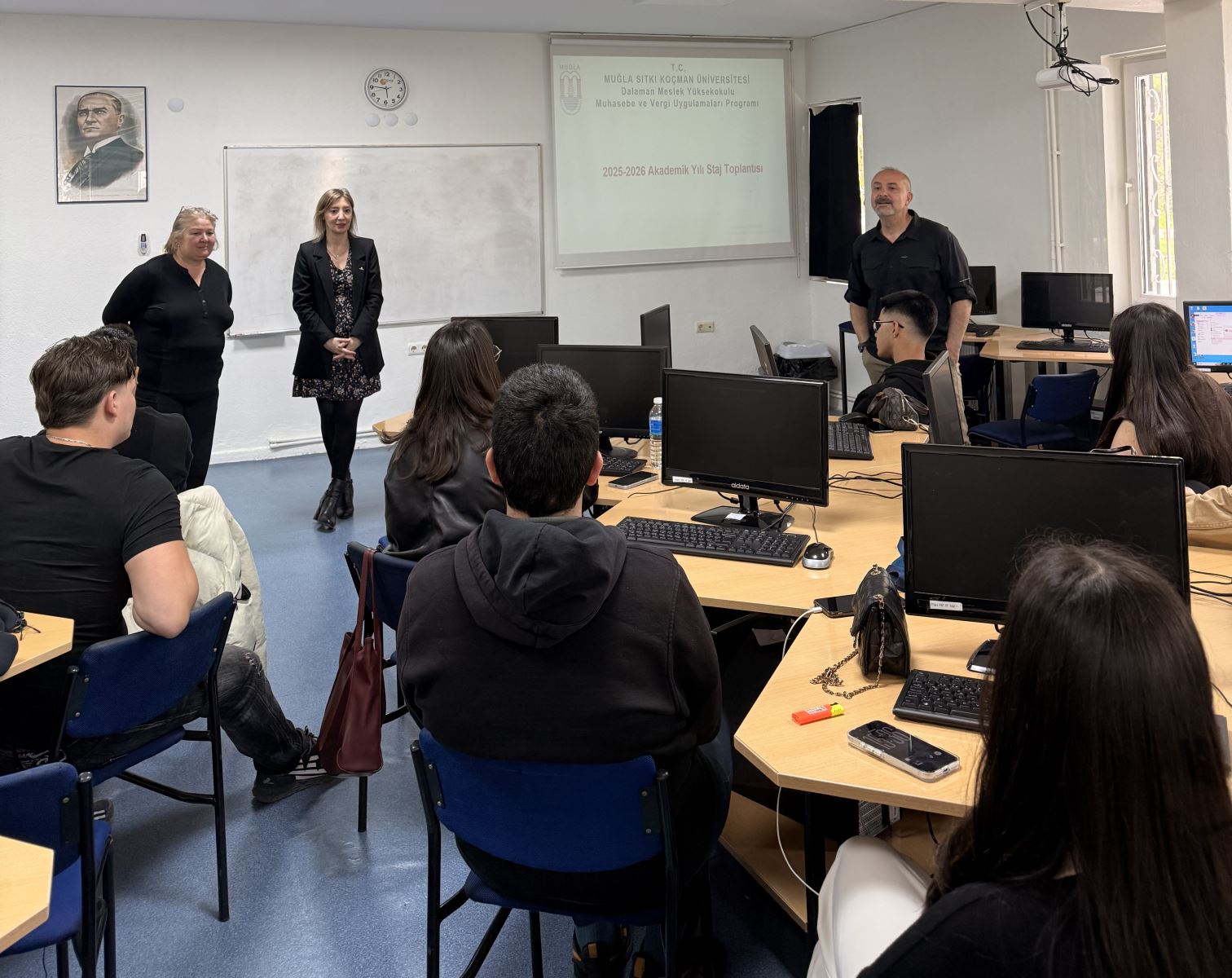 İki kadın bir erkek akademik personel Atatürk portresi önünde ve tahtada karşılarındaki 2025-2026 stajı hakkında öğrencilere bilgi veriyor. 