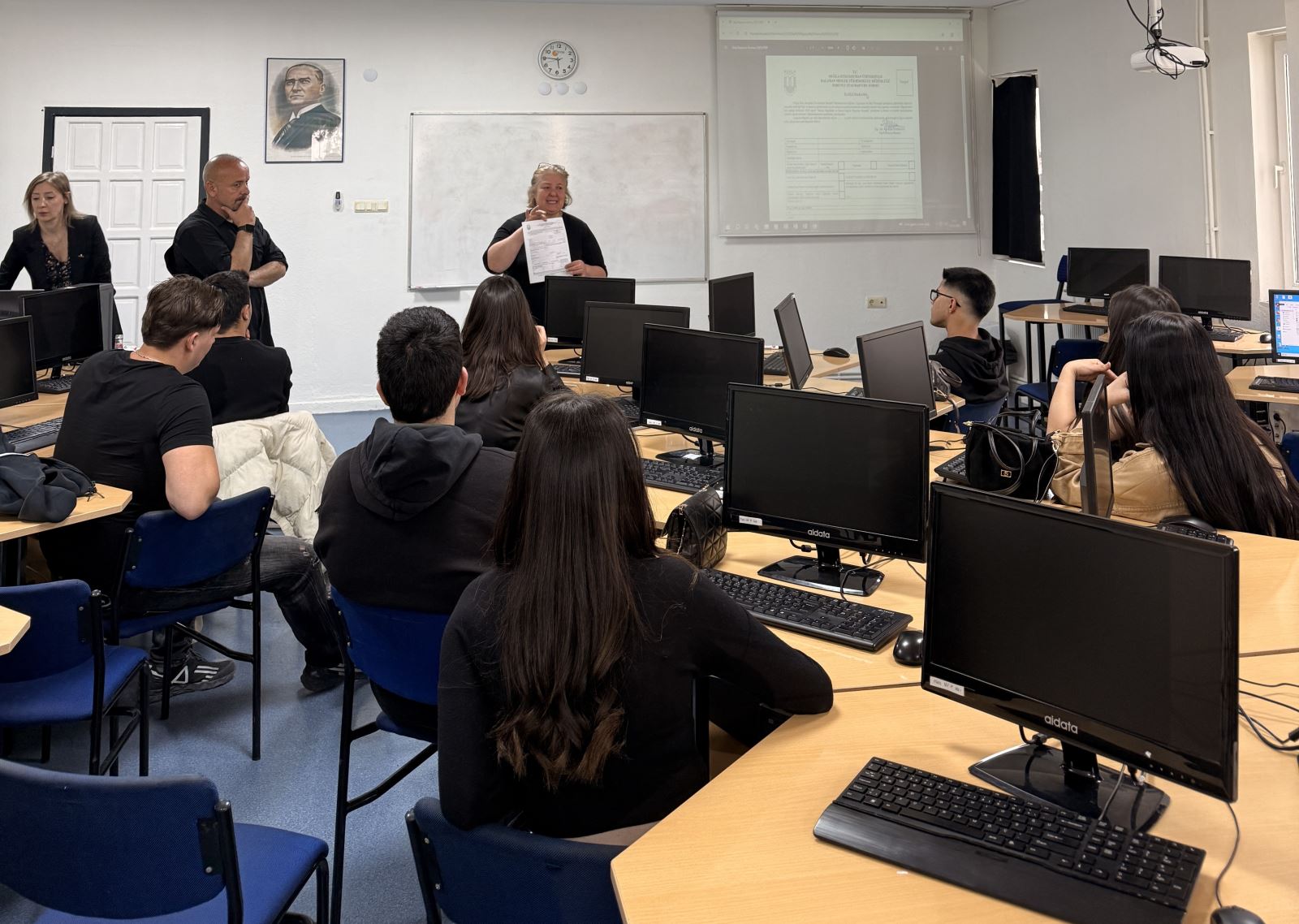 İki kadın bir erkek akademik personel Atatürk portresi önünde ve tahtada karşılarındaki 2025-2026 stajı hakkında öğrencilere bilgi veriyor. 