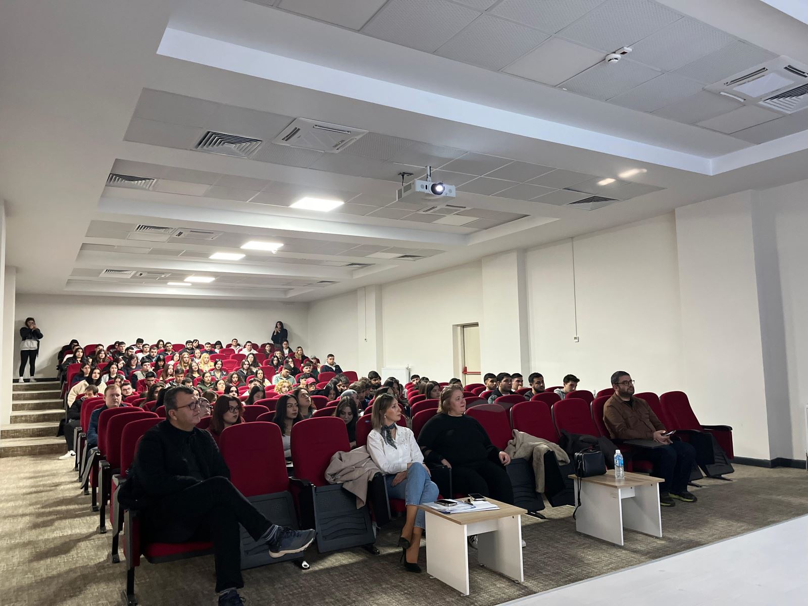 This photo shows a large conference hall filled with students seated in red chairs. In the front rows, several adults—likely instructors or speakers—are seated. The lighting is bright, and a projector hangs from the ceiling. A few people are standing against the back wall. The scene gives the impression that the event has just begun or is about to start.