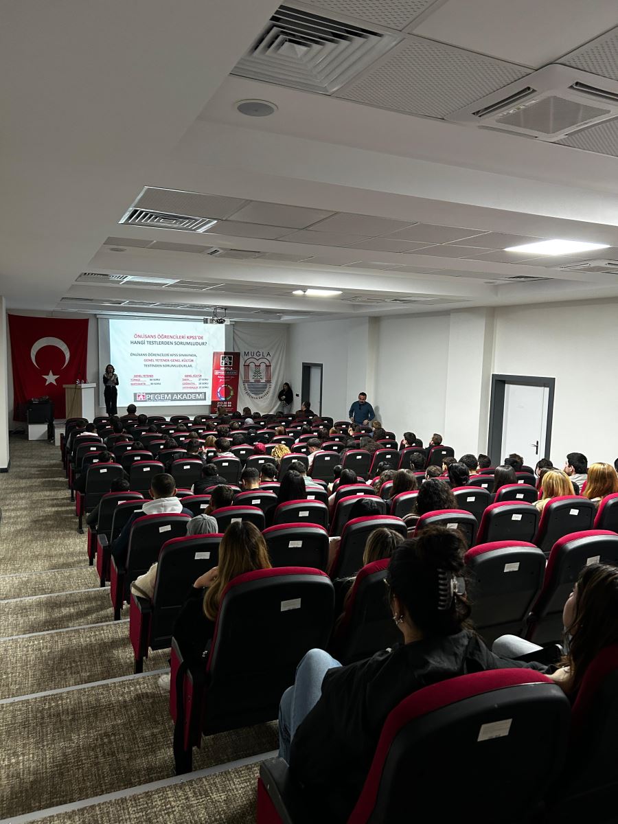 This photo is taken from the back of the hall again, showing another presentation slide on the stage. The slide reads "Önlisans Öğrencileri KPSS’de Hangi Testlerden Sorumludur?" (“Which Tests Are Associate Degree Students Responsible for in KPSS?”). Students are seated in rows, paying attention to the presentation. The hall is orderly and well-lit, and there is a speaker on stage with another person standing nearby. The Turkish flag and Muğla banner are visible behind the stage.