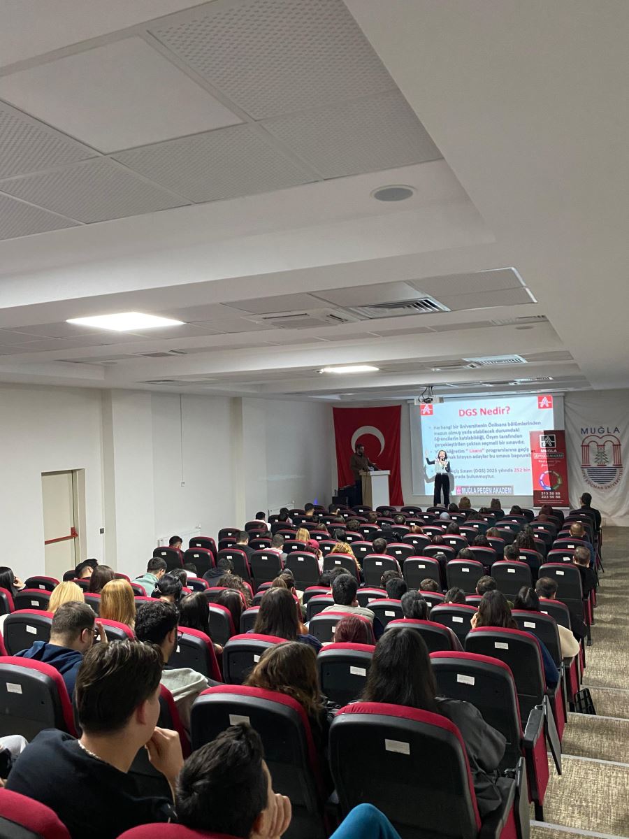 In this photo, a speaker is presenting on stage. The presentation slide displayed reads “DGS Nedir?” (“What is DGS?”). Students fill the entire hall from back to front, listening attentively. A large Turkish flag is placed on the left side of the stage, and a banner related to Muğla is on the right. Almost all seats are occupied, showing a fully engaged audience.