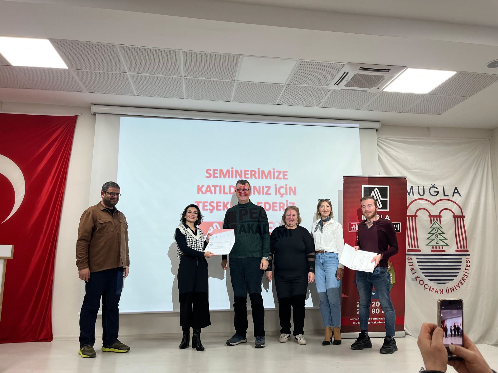 The photo captures a certificate ceremony on a stage. On the left, one man is standing, while another man in the center is receiving a certificate and posing for the photo. On the right side, four more people stand together, smiling. A projection screen behind them displays a Turkish message of thanks. A banner is visible on the right and a large Turkish flag on the left.
