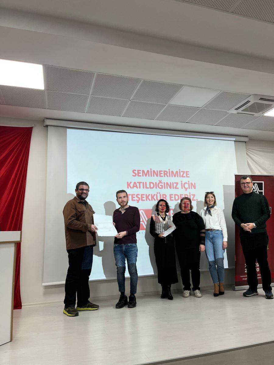 This photo also shows a certificate ceremony on stage. In the center, a woman and a tall man are posing while handing over a certificate. Other participants stand on both sides, smiling. In the bottom right corner, someone is taking a picture with a smartphone. In the background, a thank-you message, banners, and the Muğla Sıtkı Koçman University logo are visible.