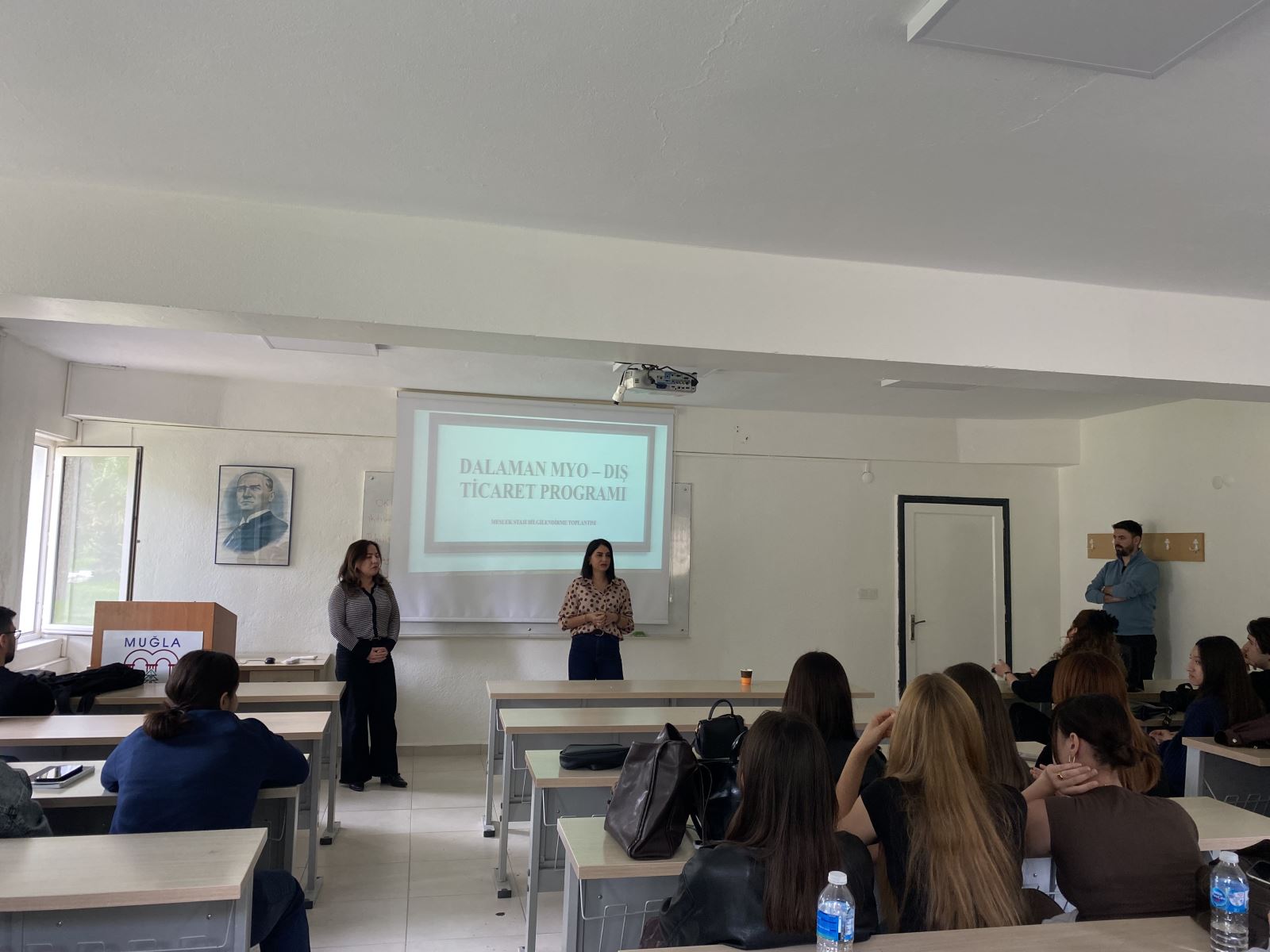 In a classroom setting, two female academic staff members are standing at the front and addressing the students. Behind them, a projected slide displays the title “Dalaman Vocational School – Foreign Trade Program.” On the left side, there is a podium and a portrait of Mustafa Kemal Atatürk on the wall. On the right side, a male lecturer stands near the wall, observing the session. Students seated in the front rows are attentively listening, some leaning on their desks. The classroom appears bright and well-organized.