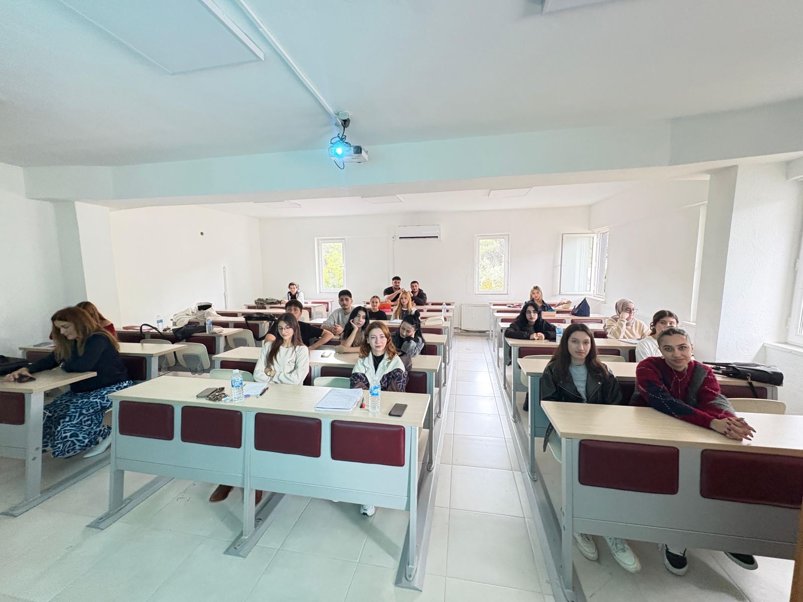 The photo was taken in a classroom setting. In a bright room with white walls, students are seated at their desks. In the front rows, a group of mostly young female students are smiling and looking toward the camera. A few male students are sitting in the back rows. On the left side of the classroom, a woman with long hair (possibly the instructor) is sitting at a desk and writing something. A projector with a blue light is mounted on the ceiling in the center of the room. Sunlight enters through the windows, creating a spacious and pleasant atmosphere. There are water bottles and notebooks on the desks. The overall mood of the photo reflects a calm moment during class