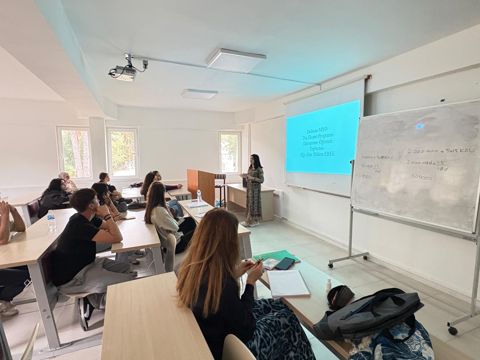 In this photo, the instructor is standing at the front of the classroom next to the whiteboard. A slide with a blue background is projected onto the screen, displaying the text: “Dalaman Vocational School Foreign Trade Program Advisor–Student Meeting Lecturer: Didem Erek.” The students are seated at their desks, attentively listening to the instructor. Some students in the front rows are taking notes in their notebooks. On the right side, there is a whiteboard with handwritten numbers and percentage signs—likely related to a classroom calculation or topic. The room is bright, with natural light coming through the windows, creating an academic and focused atmosphere