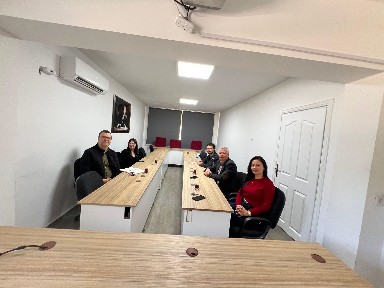Five participants are seated around U-shaped tables during a meeting in a meeting room. The wide-angle photo shows the meeting setup, a wall portrait, an air conditioner, a projector, and red chairs in the back of the room.